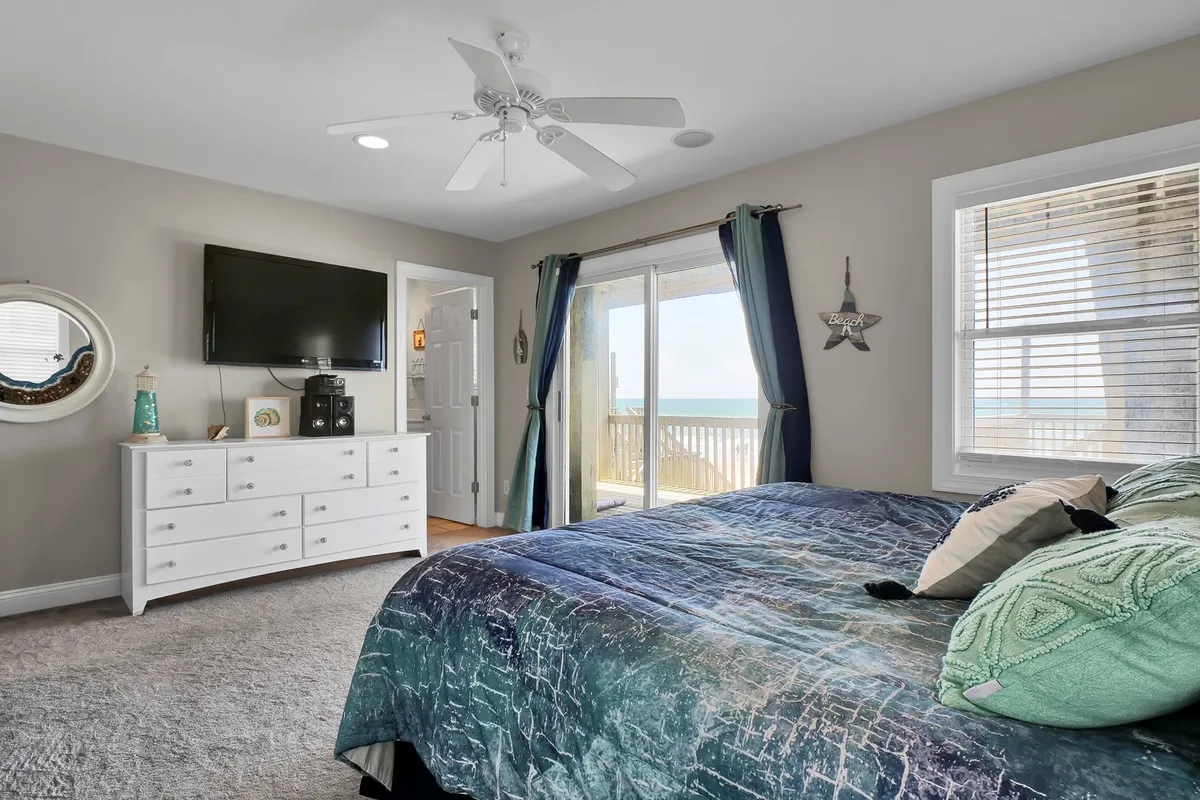 This interior image showcases a master bedroom with decorative pillows and a large window offering a direct ocean view. A dresser with a television sits beside the window, while the overall décor is tranquil and thoughtfully arranged, suggesting a space designed for relaxation and privacy.