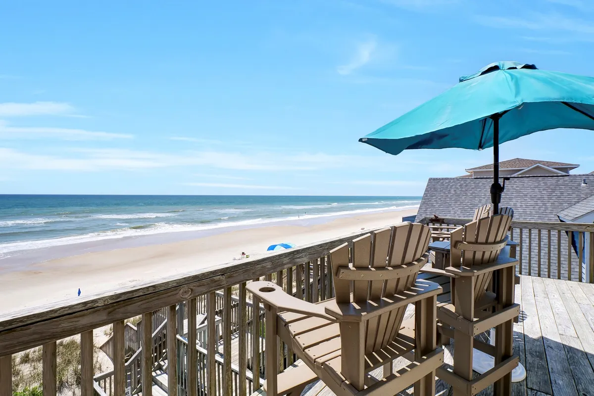 The image showcases a spacious beachfront deck overlooking the ocean, with inviting wooden deck chairs and a colorful umbrella. The scene is tranquil, with sand and waves just steps away, hinting at leisurely days spent sunbathing, reading, or socializing while enjoying breathtaking sunrise views.