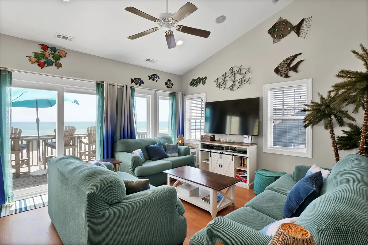 A cozy living area with large windows that frame the ocean beyond. The room features comfortable seating, colorful coastal decor, a TV unit for entertainment, and playful fish-themed wall art, creating a cheerful, welcoming atmosphere.