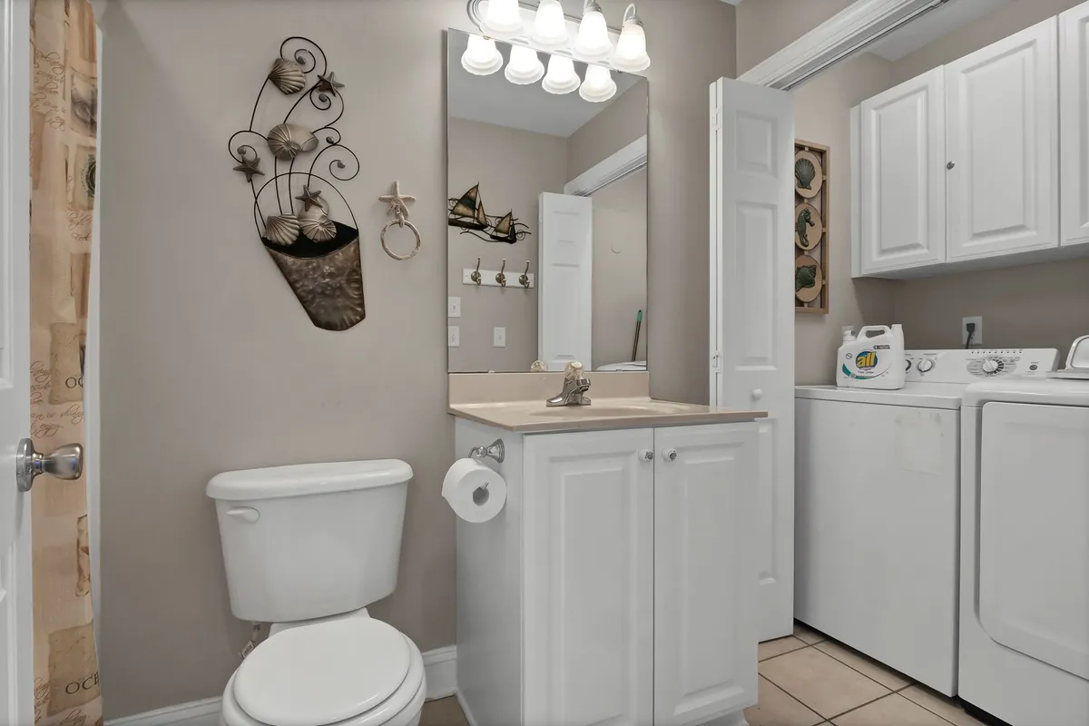 A practical bathroom and laundry area featuring a sink, mirror, toilet, washing machine, and decorative wall art. The space is tidy and thoughtfully arranged, highlighting the home's utility features.