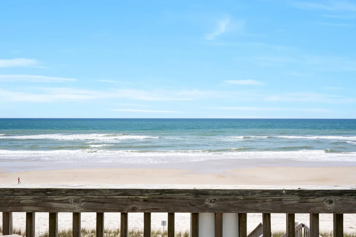 A stunning view of rolling ocean waves and a sandy beach framed by a wooden railing, with a clear blue sky overhead. The scene is open and inviting, ideal for beach walks, swimming, and soaking in breathtaking sunrises or sunsets.