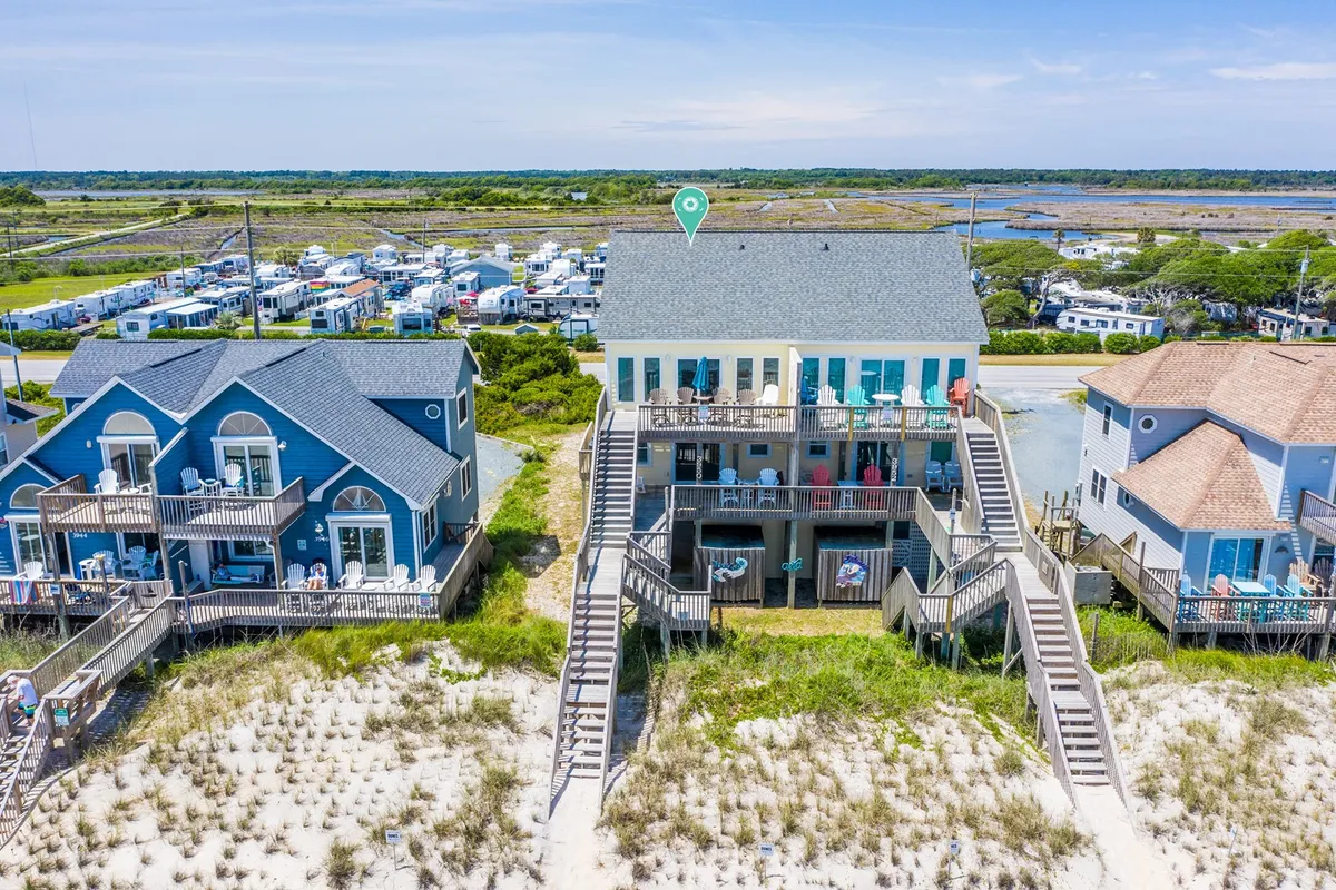 A central beach house stands just steps from the sandy pathway leading towards the shore, with adjacent homes nearby and a welcoming, cheerful vibe. The scene hints at evenings gathered on the porch and the easy access to the beach.