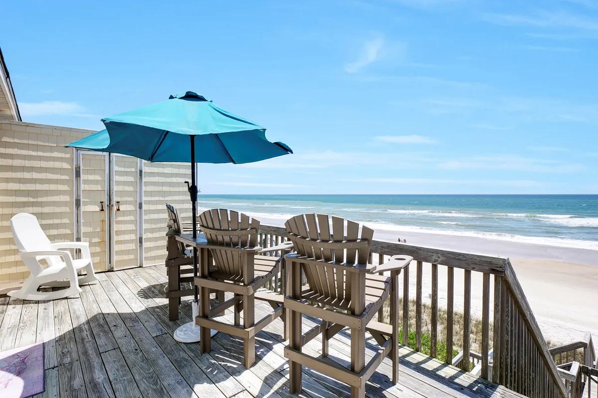 A serene beachfront deck showcases Adirondack chairs and a parasol, all overlooking the ocean. The space is designed for relaxation, socializing, and taking in stunning sunrises or sunsets with friends and family.