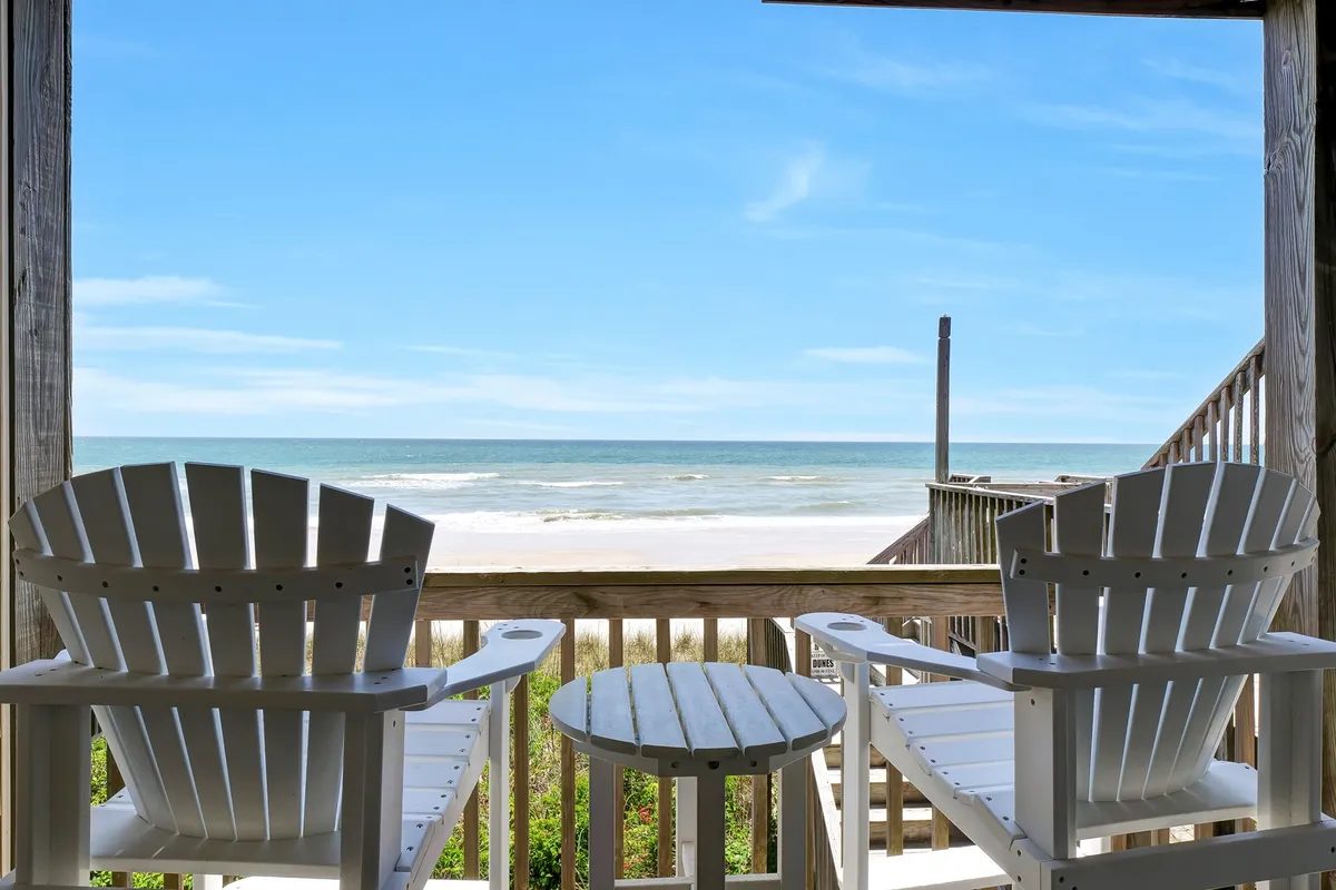 White Adirondack chairs positioned on an outdoor deck overlooking the ocean, with a table set for relaxation and conversation. The tranquil setting suggests sunrise coffee or evening stargazing, seamlessly connecting interior living to the expansive decks described in the section.