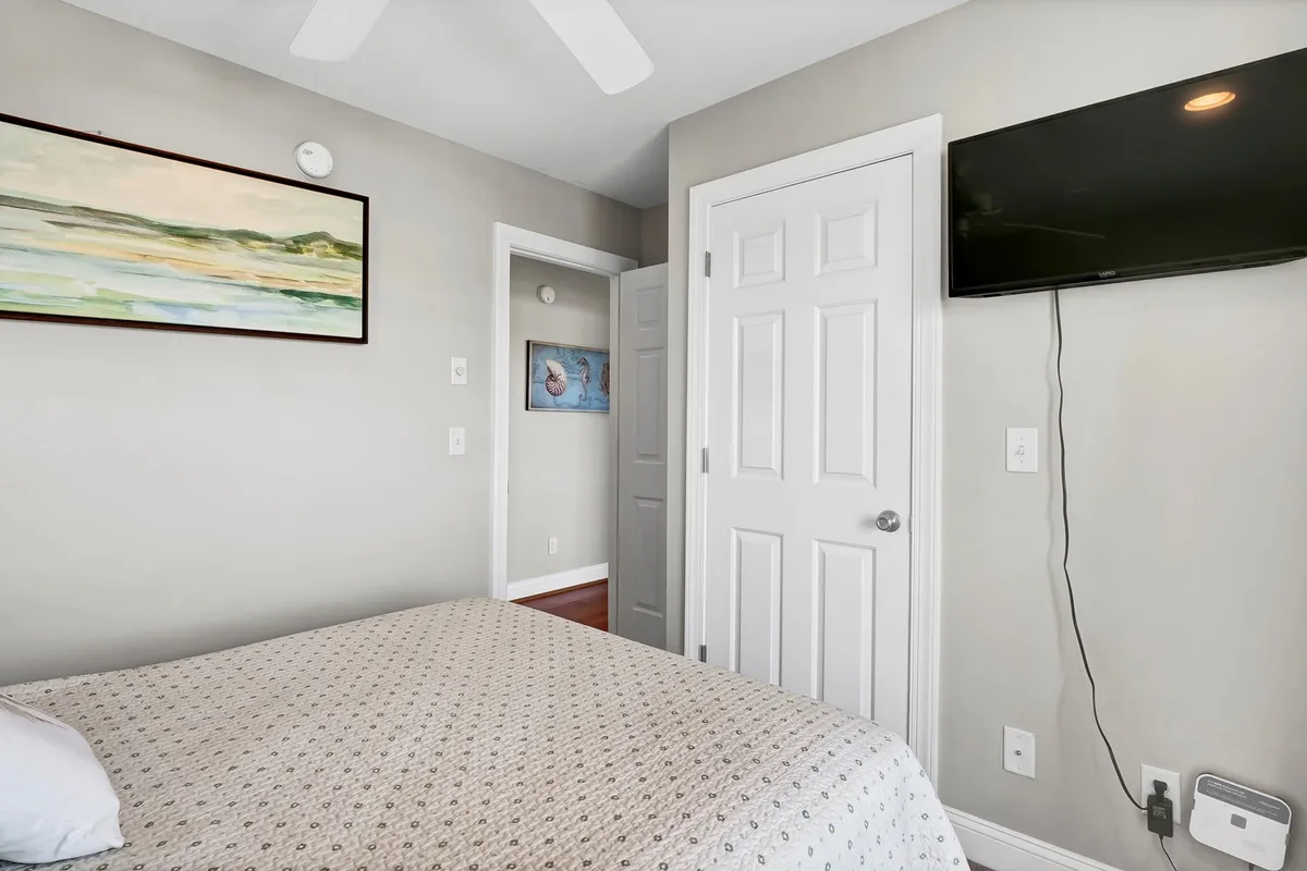 A cozy bedroom scene with patterned bedding, framed artwork, and a wall-mounted TV. The space feels serene and inviting, ideal for relaxation or unwinding after a day at the beach. The décor is bright and appealing, emphasizing comfort.