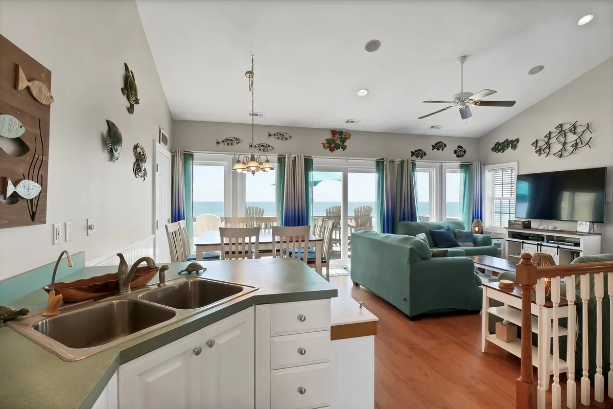 A wide-angle view capturing the open-concept living space, including the kitchen island, dining area, and living room all thoughtfully arranged to maximize the stunning ocean view visible through large windows. The space features comfortable seating, a central dining table, and a seamless flow perfect for gatherings.