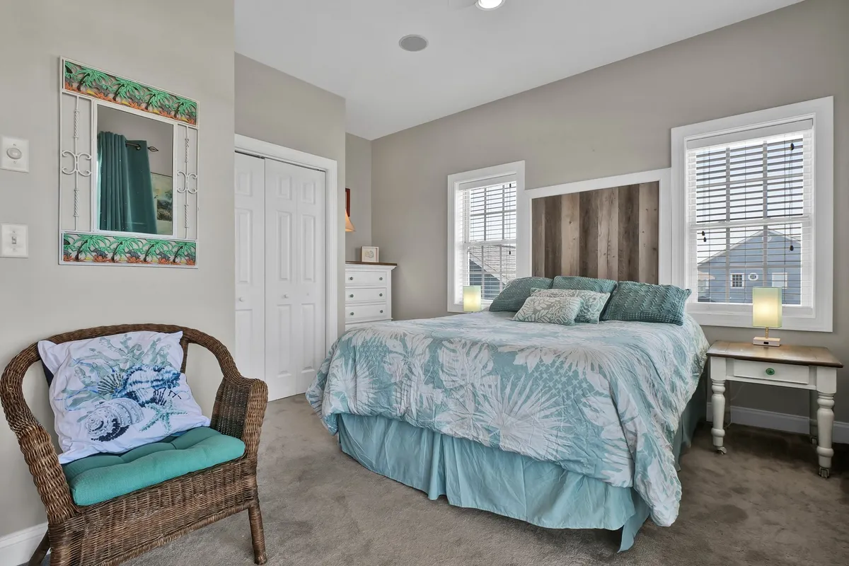 A tranquil and cozy bedroom featuring a bed with decorative bedding, a wicker chair, and a framed mirror, all bathed in gentle natural light. The intimate arrangement suggests a space perfect for reading, relaxing, or sharing quiet moments.