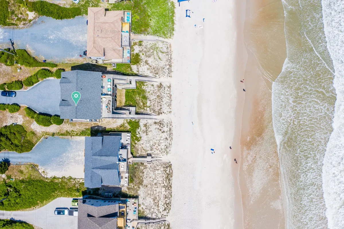 Families and friends are seen enjoying the expansive sandy beach in front of beachfront homes. The wide ocean, gentle surf, and a lively but relaxed atmosphere set the stage for sun-filled days spent playing, swimming, and relaxing together.