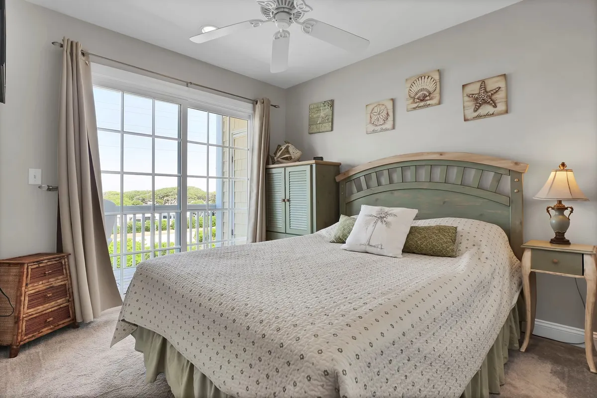 A bright and tranquil bedroom featuring a comfortable bed, a large window that lets in ample natural light, decorative wall art, and a cozy nightstand—ideal for relaxing after a day at the beach.